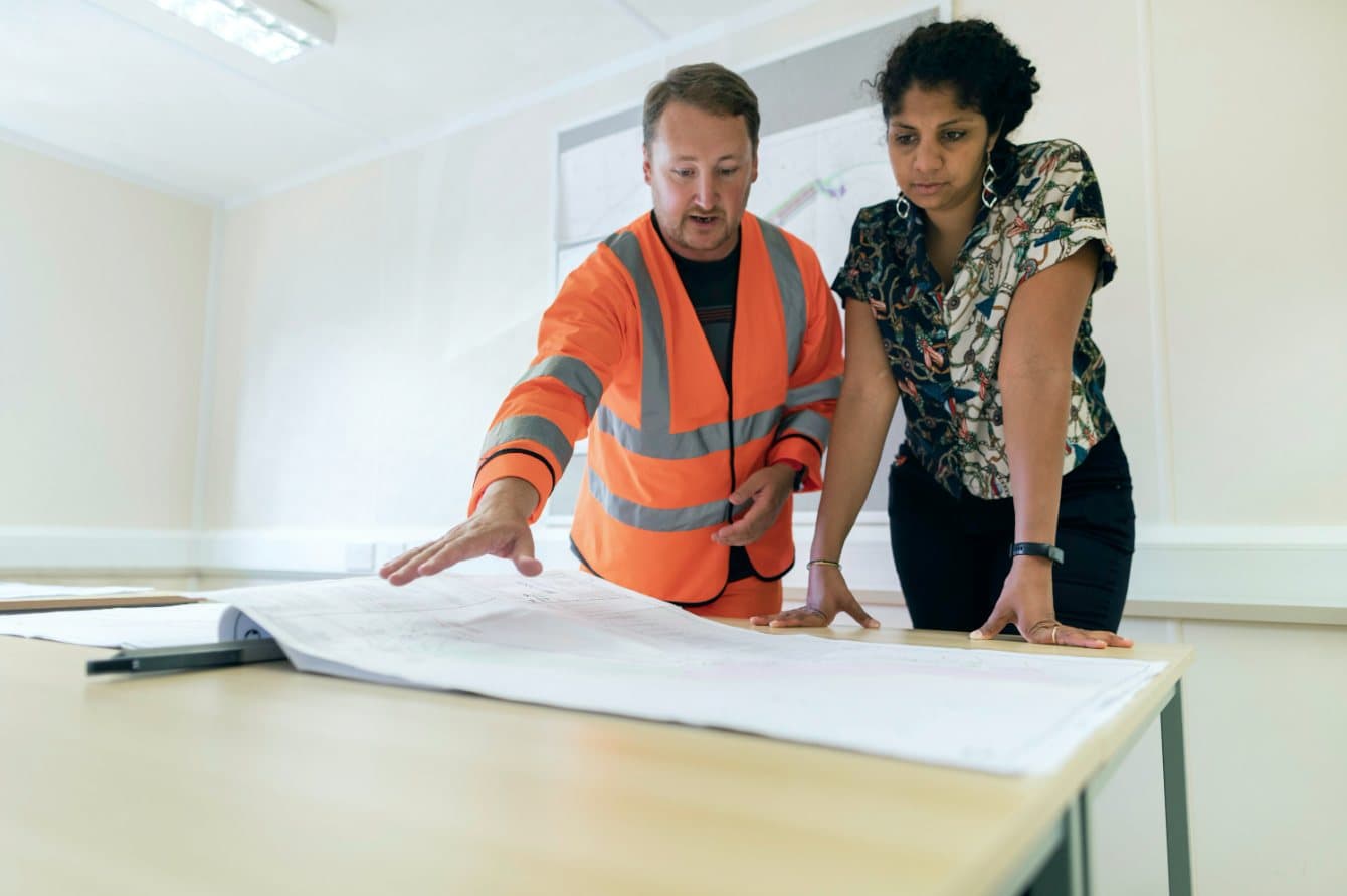 A site engineer in a high-visibility vest and a colleague reviewing plans on a table.